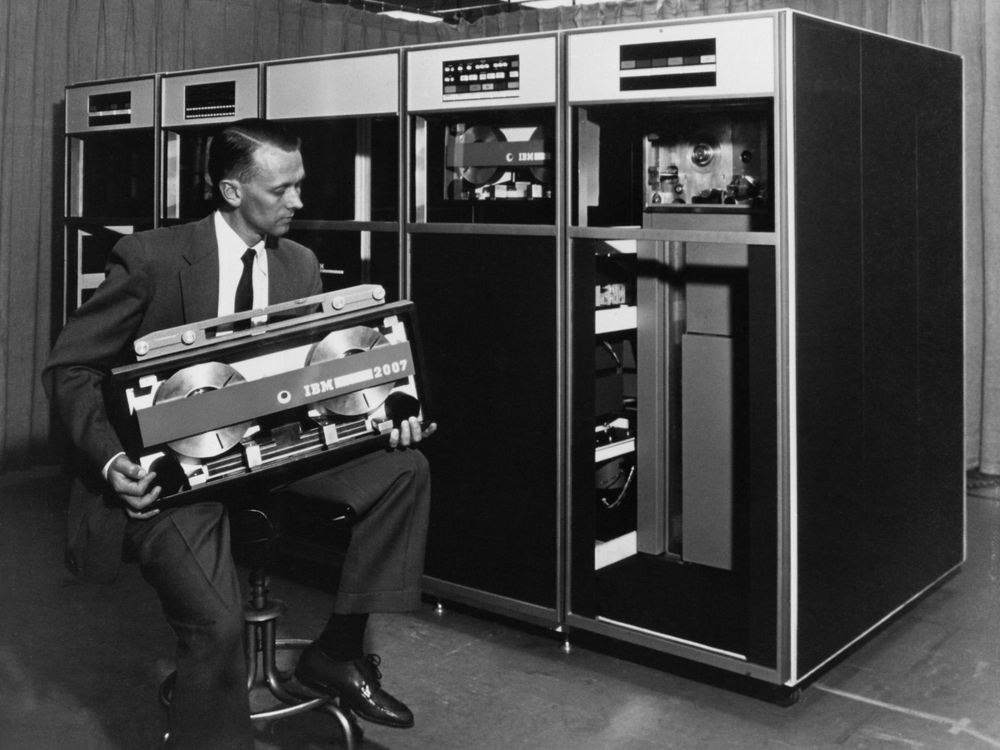 Техник IBM держит одну из массивных кассет данных перед шкафами ленточных накопителей. Такие кассеты использовала автоматизированная система Tractor компьютера Harvest. / IBM.